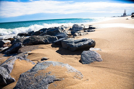 Sunny beach at the day with wet rocks, white clouds and small buildings in the backgroundの写真素材