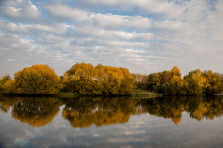 Autumn forest with reflection in the pond river. Sky with clouds.