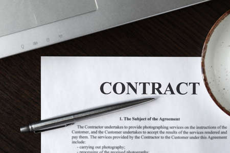 Cup of coffee with foam, silver ballpoint pen, blank contract and laptop on a wooden table