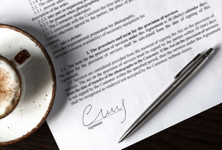 Contract signing concept. Close-up signature with silver pen and cup of coffee on dark wooden table...