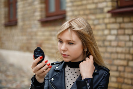 A young girl wearing black jacket, checking how she looks in small portable mirror, bricks in background.の写真素材