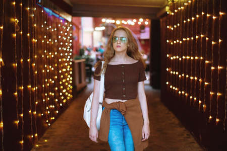 Beautiful girl in a green glasses walks in the evening city with garlands and light bulbs.の写真素材