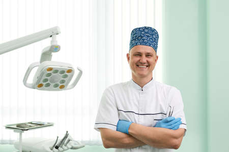 Highly qualified male dentist standing at his clinic smiling to the camera with his arms crossed near modern dental equipment.の写真素材