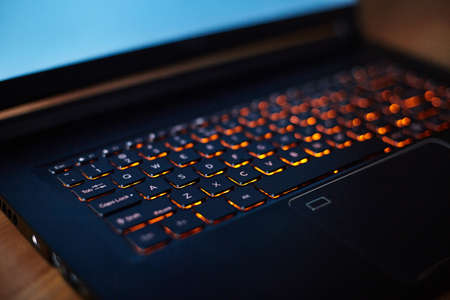 Laptop keyboard with orange neon illumination, backlit keyboard. Black laptop with blue monitor.