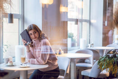Beautiful young woman reading a book with a cup of cappuccino sitting in the cafe.の写真素材