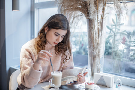 Portrait of beautiful woman reading a book while relaxing in the cafe. Cappuccino and cake are on the table.の写真素材