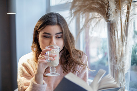 Beautiful young woman reading a book with a cup of cappuccino sitting in the cafe.の写真素材