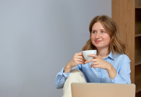 Beautiful young woman working on laptop computer while sitting at the living room, drinking coffee.の写真素材