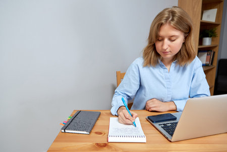 Young happy woman sitting at desk, working on computer at home. Pleasant attractive smiling lady looking at laptop screen, shopping, chatting in social networks, studying online remotely.の写真素材