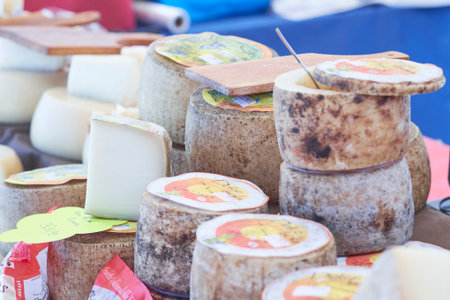 Stacks of artisanal cheeses are showcased on a vendor's table at a bustling market. Bright labels and variety highlight the craftsmanship and flavors of each cheese available.の写真素材