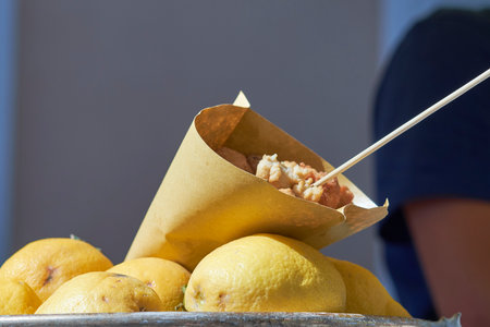 A vendor offers crispy fried food on a stick, presented in a paper cone resting on a mound of fresh lemons at a bustling outdoor market. Bright sunlight enhances the vibrant colors.の写真素材
