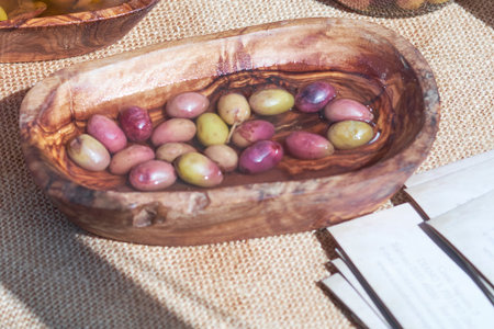 Various olives float in a rustic wooden bowl, placed on a natural fabric surface. Nearby, informational cards await visitors at the bustling local market in the bright afternoon light.の写真素材