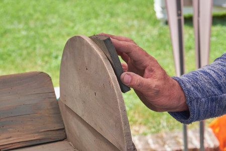 A close-up of a hand with a sanding block smoothing the rounded edge of a light-colored wooden furniture piece, likely part of a restoration project, with a blurred green background.の写真素材
