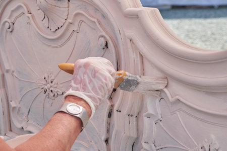 A close-up view of a gloved hand using a paintbrush to meticulously apply white paint to the intricate, ornate details of a piece of furniture, highlighting the process of restoration or DIY work.の写真素材