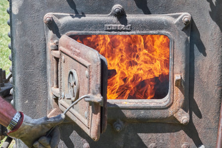 A gloved hand pulls open the heavy, rusted cast iron door of an industrial furnace, revealing a...