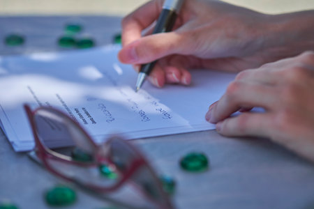 A close-up shows a person's hands holding a pen and writing on a white paper document, likely a...