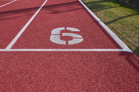 Detail of lane number 6 on a red outdoor running track beside grass, symbolizing athleticism, focus, and the starting point of a race or journey.の写真素材