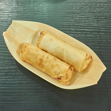 Two golden crispy spring rolls served in an eco-friendly bamboo leaf tray, presented on a dark...