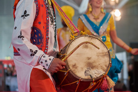 Close-up of a man playing a traditional Punjabi dhol drum during a vibrant Bhangra dance performance in colorful ethnic attire at a cultural festival.の写真素材