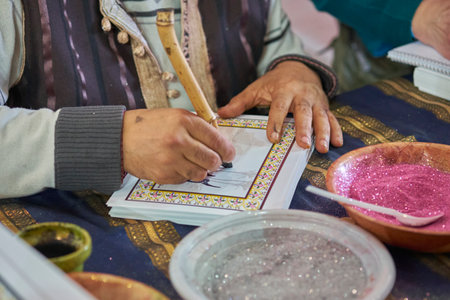 Close-up of an artisan creating traditional calligraphy with a bamboo pen, surrounded by colorful glitter and ink bowls on a patterned fabric table.の写真素材