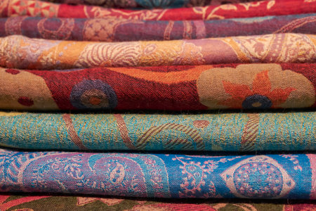 A close-up view of stacked, colorful traditional fabrics with intricate patterns, showing texture,...