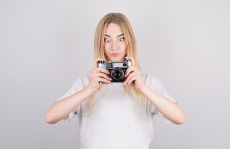 Expressive portrait of a beautiful young surprised blonde woman in a white T-shirt in the studio on a gray background with a camera in her handsの写真素材