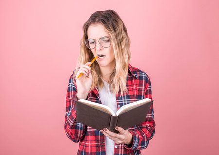 Portrait of thoughtful pensive girl having notebook and pencil in mouth. Isolated on pink background...