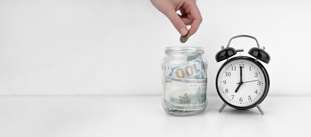 Mans hand put coin into a glass jar with savings near black alarm clockの写真素材