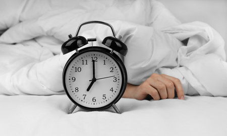 Woman's hand lying near black alarm clock on bedの写真素材