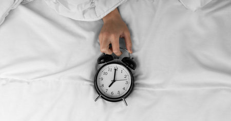 Woman's hand holding black alarm clock on bedの写真素材