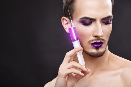 Pretty sensual gay man with art makeup and beard. Color syringe in the hand.の写真素材