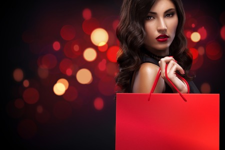 Beautiful young woman make shopping in black friday holiday. Girl with red bag on background with christmas lights.の写真素材