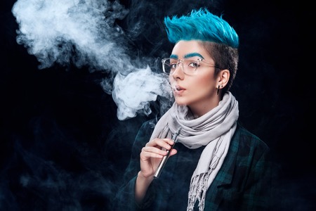 hipster woman with blue hair smoking vape on black backgroundの写真素材