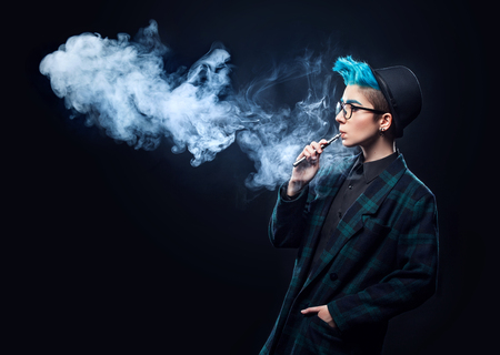 hipster woman with blue hair smoking vape on black background. Copy spaceの写真素材