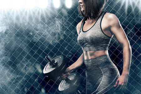 Brutal athletic woman pumping up muscles with dumbbells on black background with smokeの写真素材