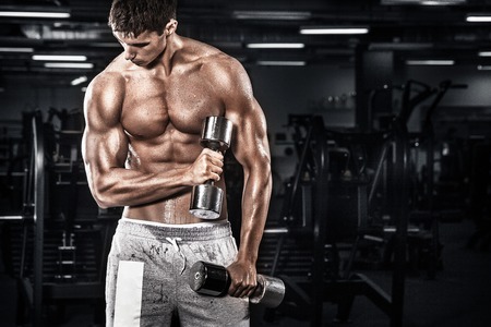 Athletic shirtless young sports man - fitness model holds the dumbbell in gym. Copy space fore your text.の写真素材