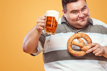 Young fat man at oktoberfest, drinking beer and eating pretzel on yellow background.の写真素材
