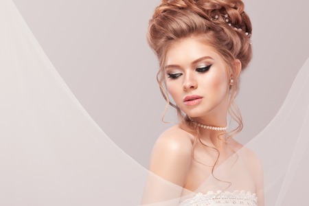 Beautiful bride with fashion wedding hairstyle - on gray background. Closeup portrait of young gorgeous bride. Wedding. Studio shot with copy space. Beautiful bride portrait with veil over her faceの写真素材