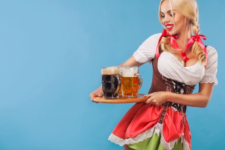 Young sexy Oktoberfest girl - waitress, wearing a traditional Bavarian dress, serving big beer mugs on blue background.の写真素材
