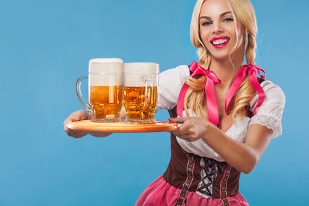 Young sexy Oktoberfest girl - waitress, wearing a traditional Bavarian dress, serving big beer mugs on blue background.の写真素材