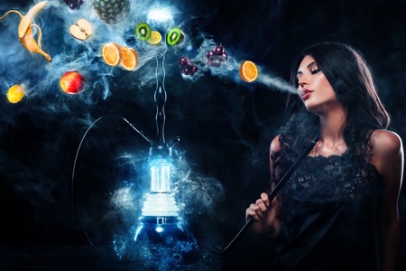 Young, beautiful woman in the night club, bar smoke a hookah or shisha. The pleasure of smoking. Fruits in the smoke. Copy space. Hookah advertisement concept.の写真素材