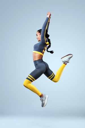 Attractive excited fitness girl dancer in sportwear jumping of joy isolated over gray backgroundの写真素材