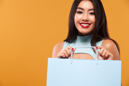 Happy asian woman at shopping holding blue bag isolated on orange background on black friday holiday. Copy space for sale ads.の写真素材