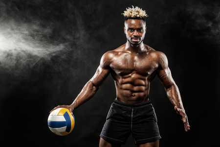 Portrait of afro-american sportsman, valleyball player with a ball over black background. Fit young man in sportswear holding ball.の写真素材