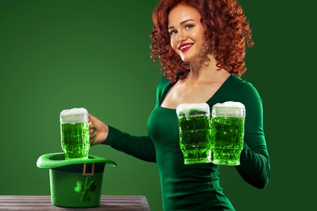 Saint Patricks Day. Young ginger Oktoberfest leprechaun, wearing grenn dress, serving big beer mugs on green background with copy spaceの写真素材