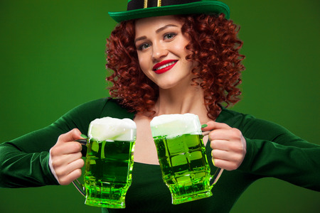 Saint Patricks Day. Young ginger Oktoberfest leprechaun, wearing grenn dress, serving big beer mugs on green background with copy spaceの写真素材