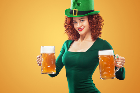 Saint Patricks Day. Young ginger Oktoberfest leprechaun, wearing grenn dress and hat, serving big beer mugs on orange background..の写真素材