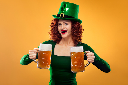 Saint Patricks Day. Young ginger Oktoberfest leprechaun, wearing green dress and hat, serving big beer mugs on orange background..の写真素材