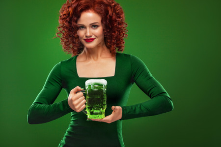 St Patricks Day. Young sexy and redhead Octoberfest waitress, wearing a dress, serving big beer mugs on green background with copy spaceの写真素材