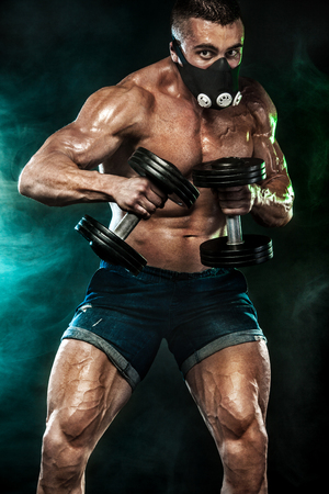 Muscular young fitness sports man in training mask. Workout with dumbbell in fitness gymの写真素材